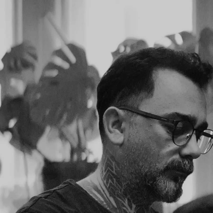 Erkan, black and grey realism specialist, at his bench by the studio window, glasses on, neck and throat tattooed.