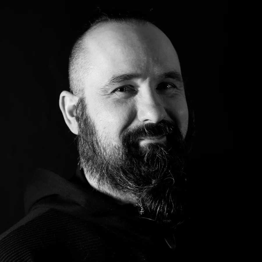 Kris, neo-traditional specialist, photographed against black in the studio, shaved head and full beard, black hooded sweatshirt.