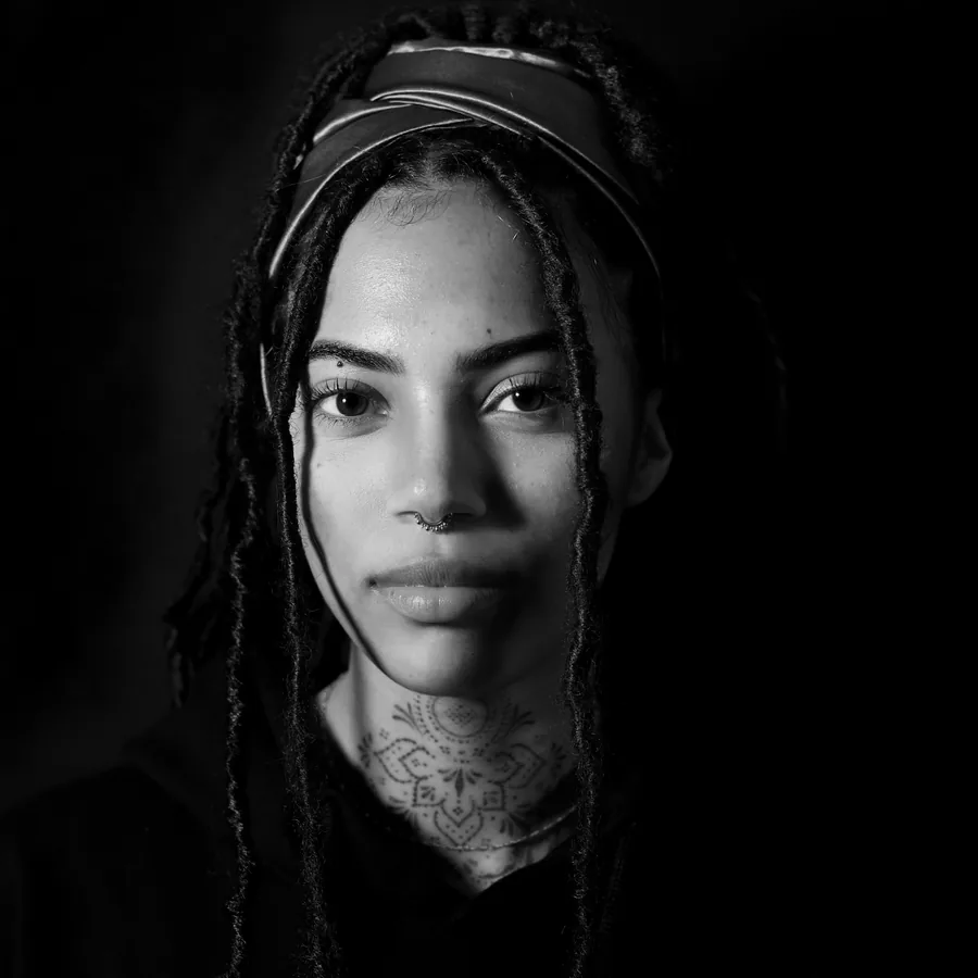 Renee, fine line specialist, photographed against black in the studio, locs tied back, script tattoo across the collarbone.