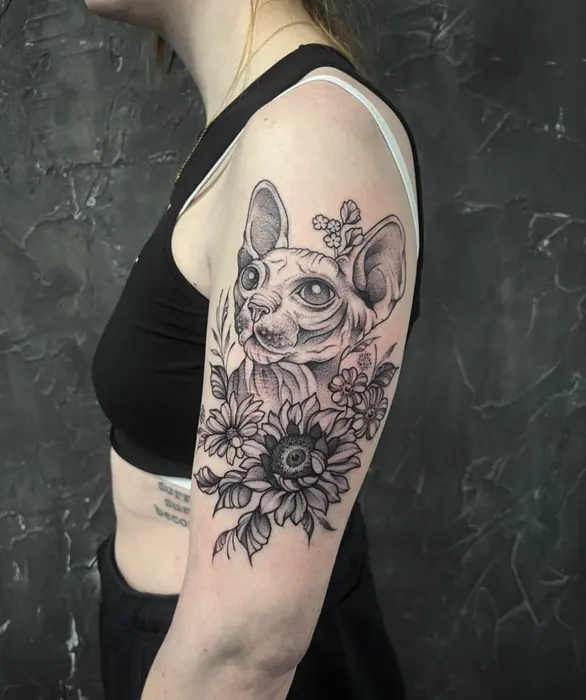 A fine line upper-arm piece: a sphynx cat framed by botanical linework.