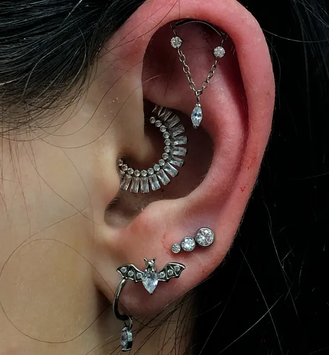 A conch piercing with a fine chain running to the upper helix, with a bat-shaped lobe stud beneath.