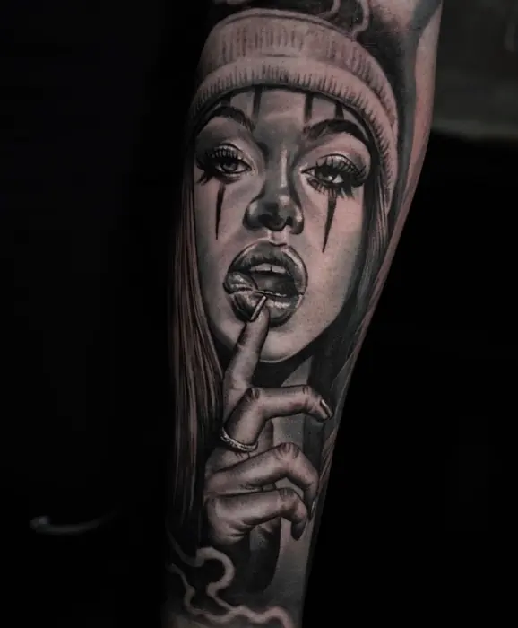 A black and grey realism portrait on the forearm: a woman in a beanie, finger raised to her lips, tear detail under both eyes.